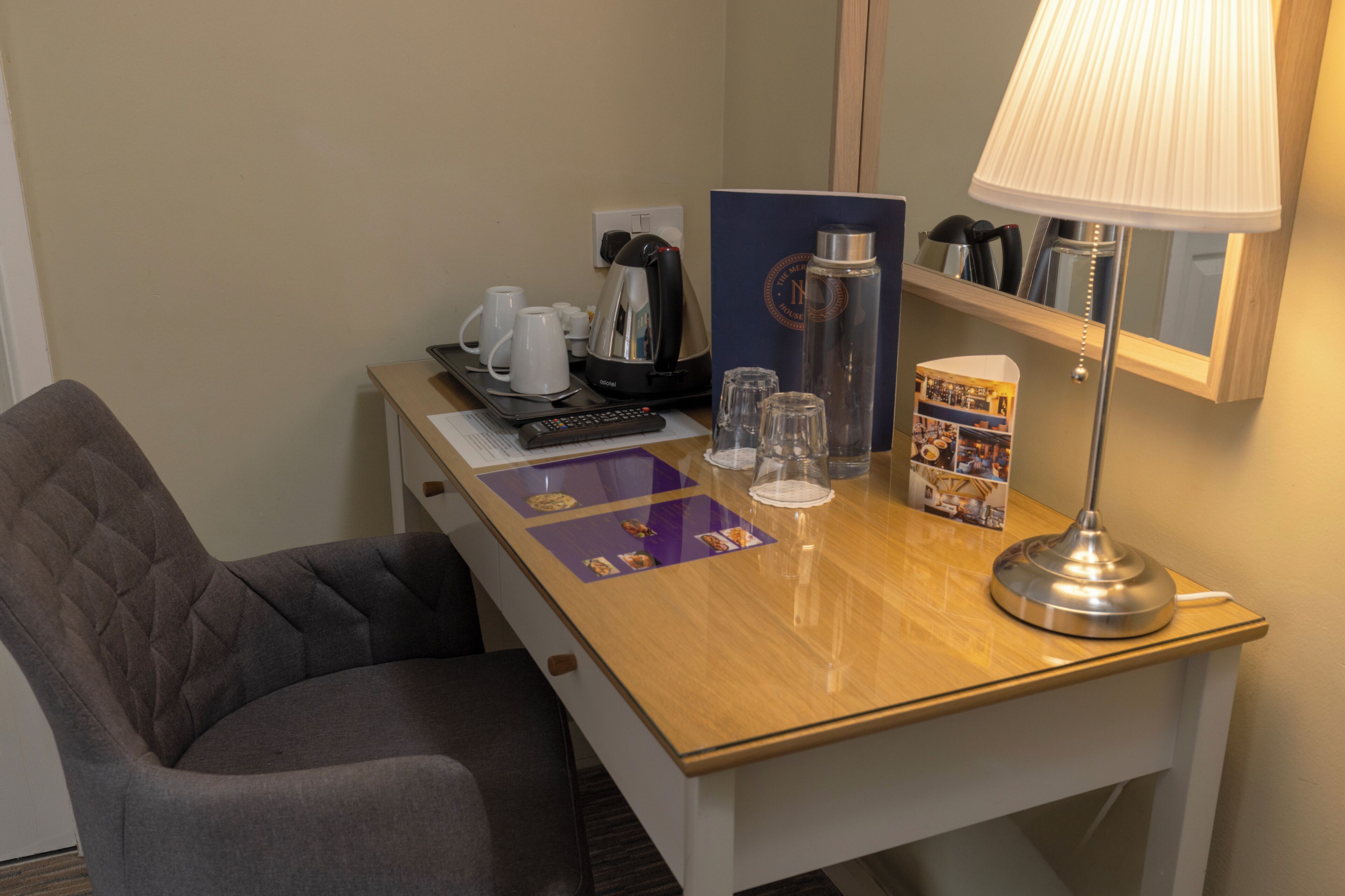 A hotel room with a wooden desk, a grey upholstered chair, a remote control, a coffee maker, a water bottle, a lamp, and a magazine.