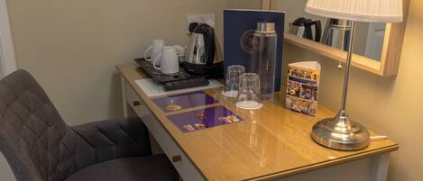 A hotel room with a wooden desk, a grey upholstered chair, a remote control, a coffee maker, a water bottle, a lamp, and a magazine.