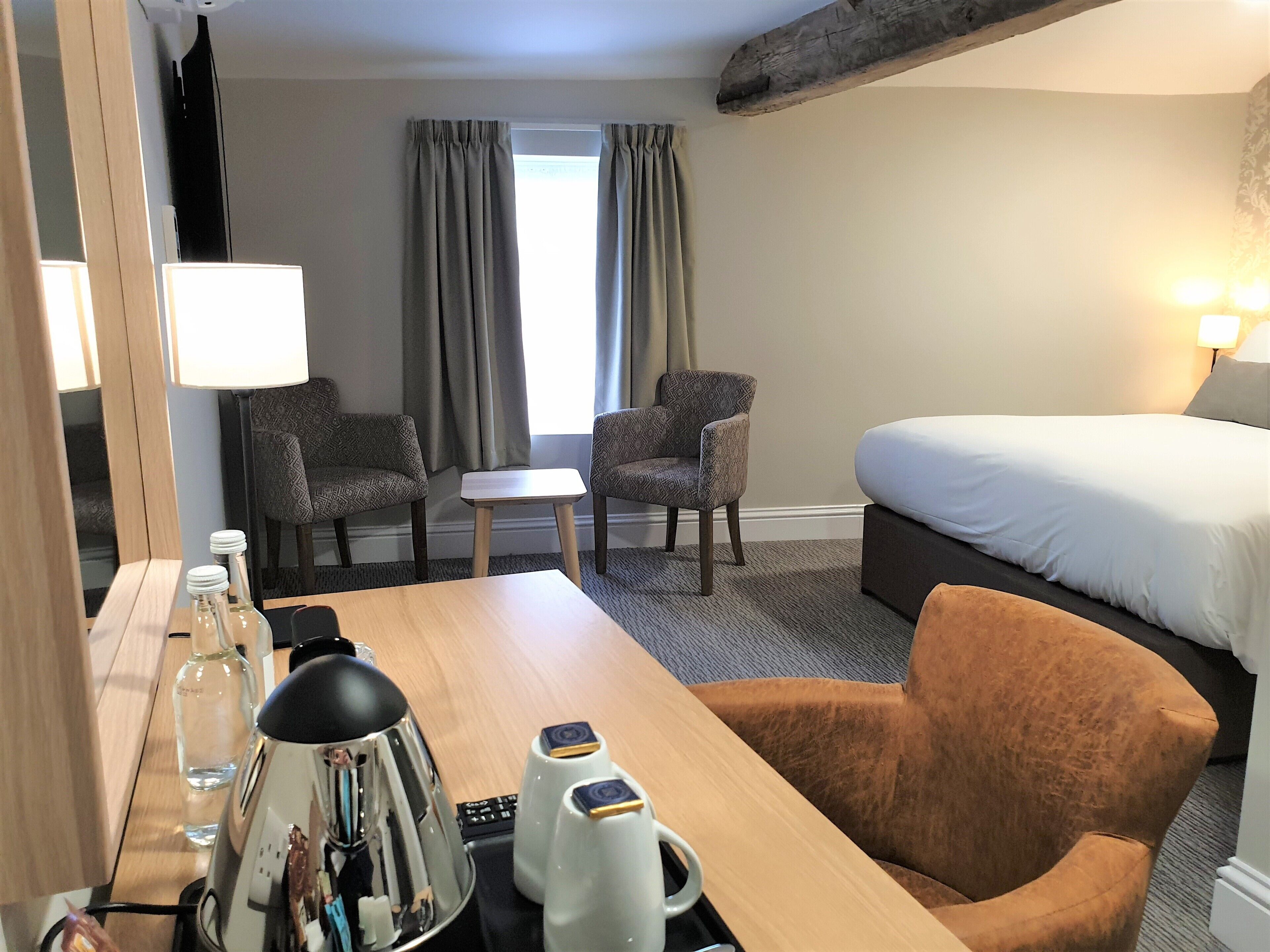 A hotel room with a bed, two armchairs, a small table, a desk with a lamp, a mirror, and a coffee maker.