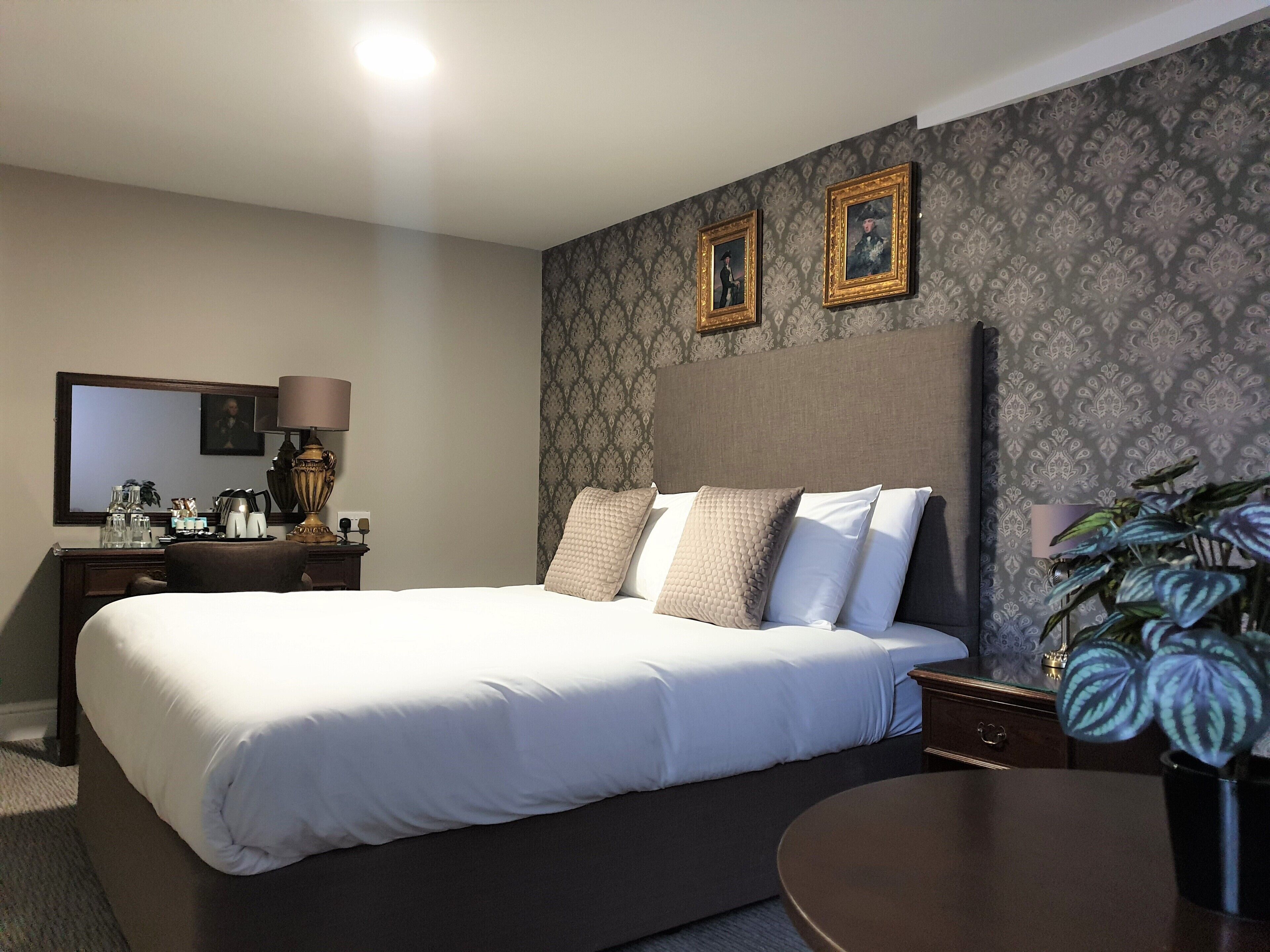 Ein Hotelzimmer mit einem großen Bett, einem Nachttisch mit Lampe, einem Schminktisch und einem Spiegel. Über dem Bett befinden sich zwei gerahmte Bilder an der Wand.