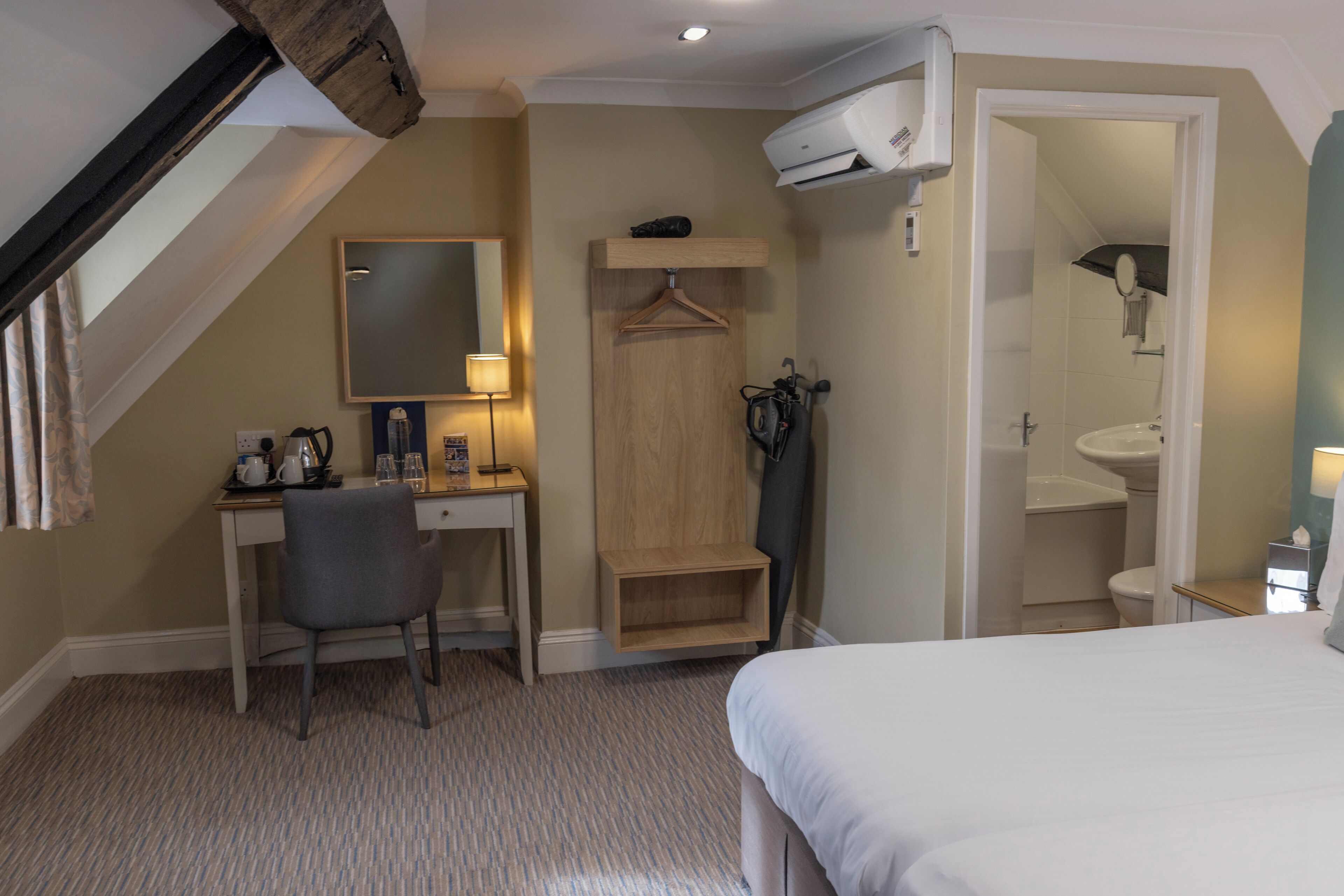 A hotel room with a bed, a desk with a chair, a hanging coat rack, a mirror, and a bathroom with a sink and toilet.
