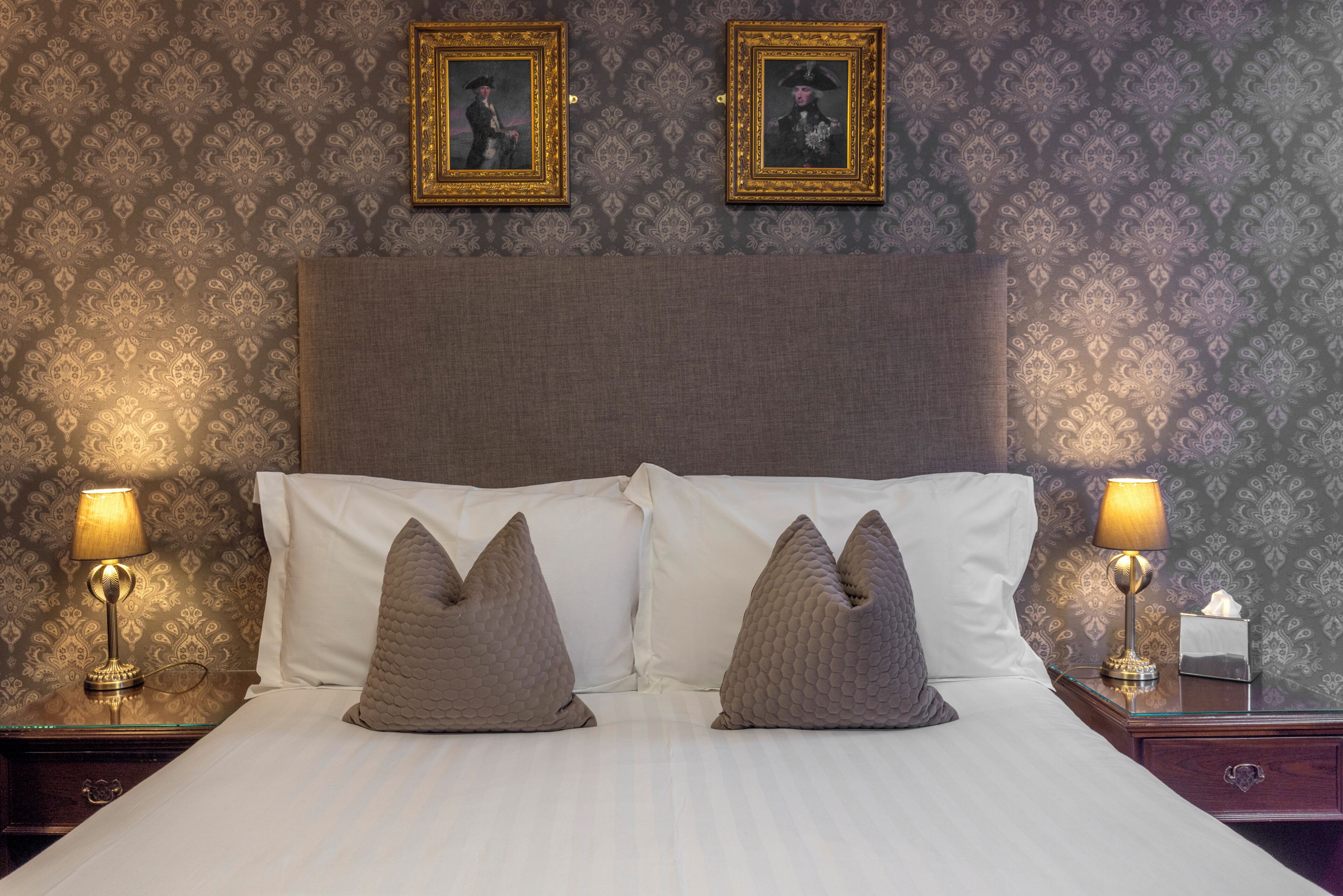 A neatly made bed with two pillows, a headboard, two framed pictures on the wall, and two bedside lamps.
