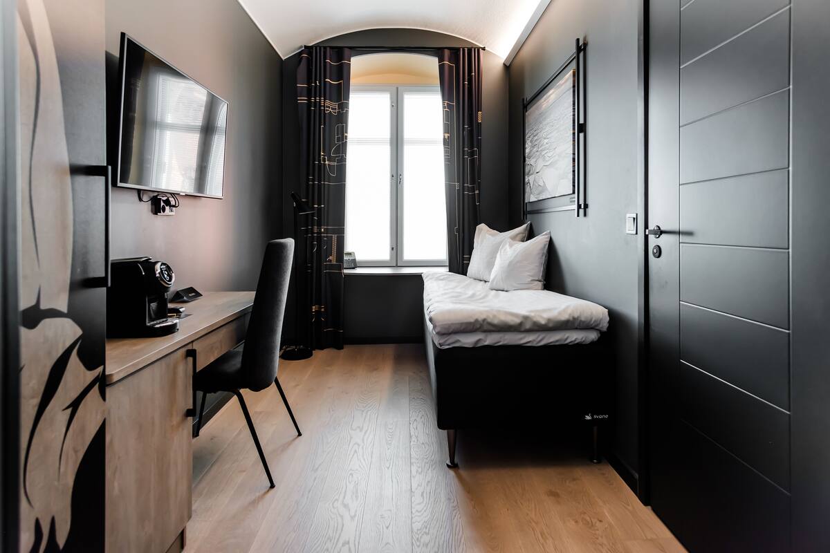 A compact hotel room with a bed, a desk, a chair, a TV, and a window with curtains.