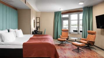 Quarto de hotel com uma cama, duas poltronas, uma mesinha, televisão e janela com cortinas.