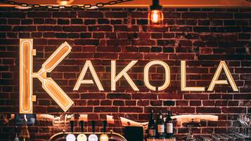 A bar with a neon sign spelling "KAKOLA" on a brick wall, bar equipment, and glasses.