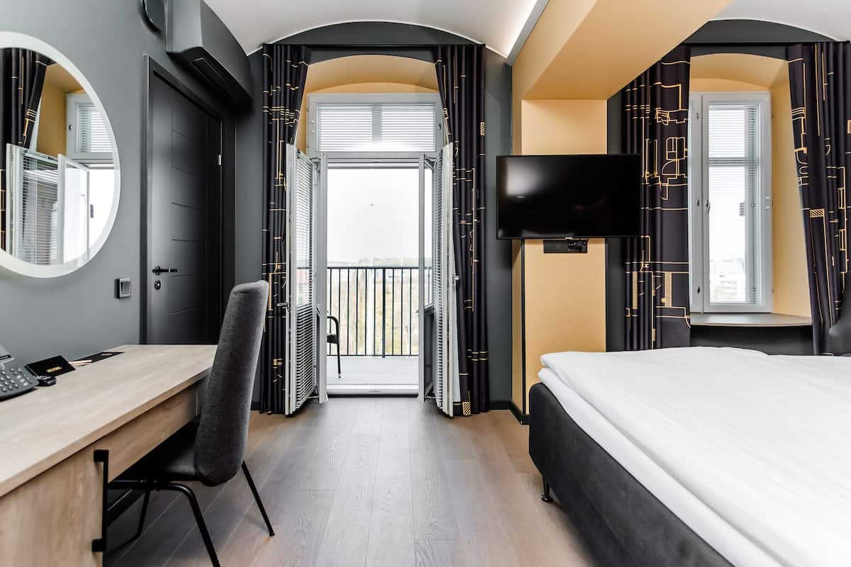A hotel room with a bed, a desk, a chair, a TV, and a balcony with curtains.
