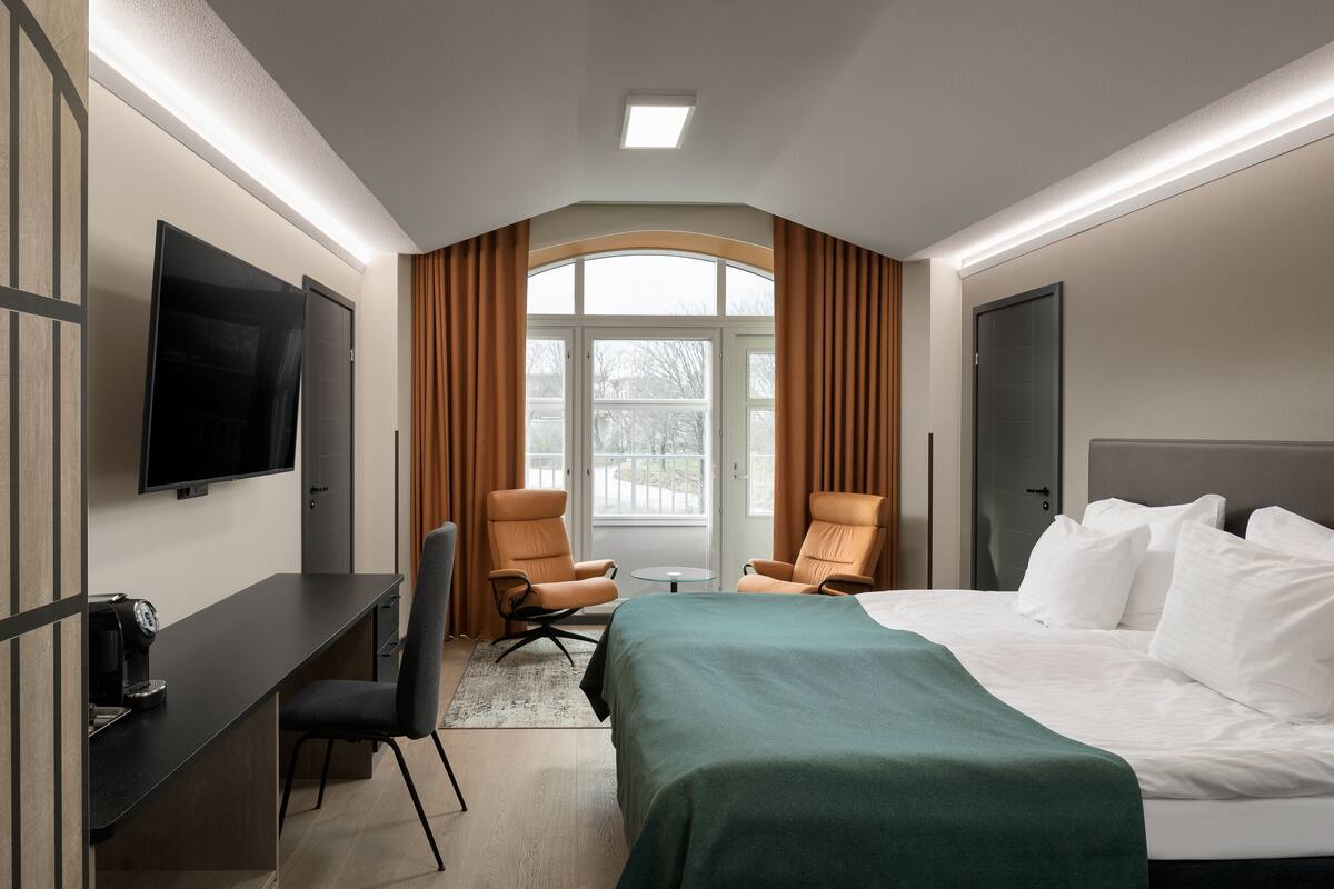A hotel room with a bed, two armchairs, a desk with a TV, and a large window with curtains.