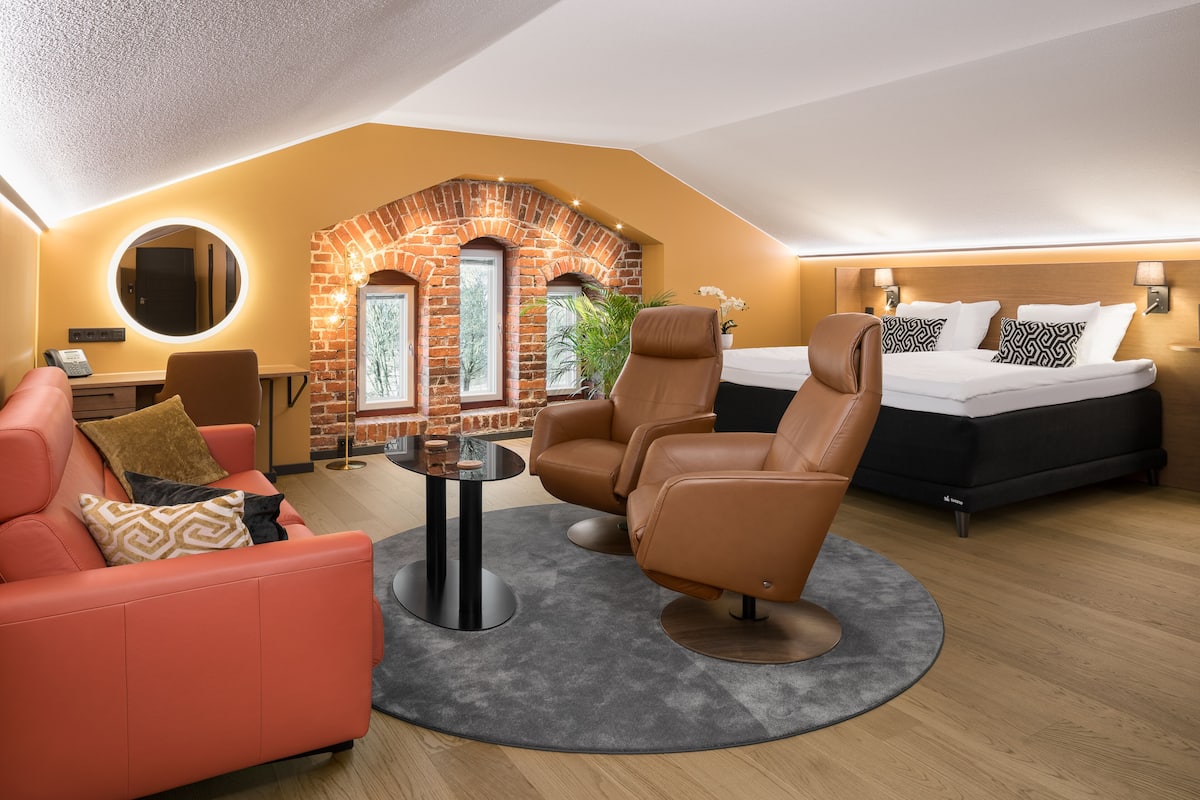 A modern hotel room with a brick archway, a round mirror, a sofa, a recliner, a bed, and a small table.