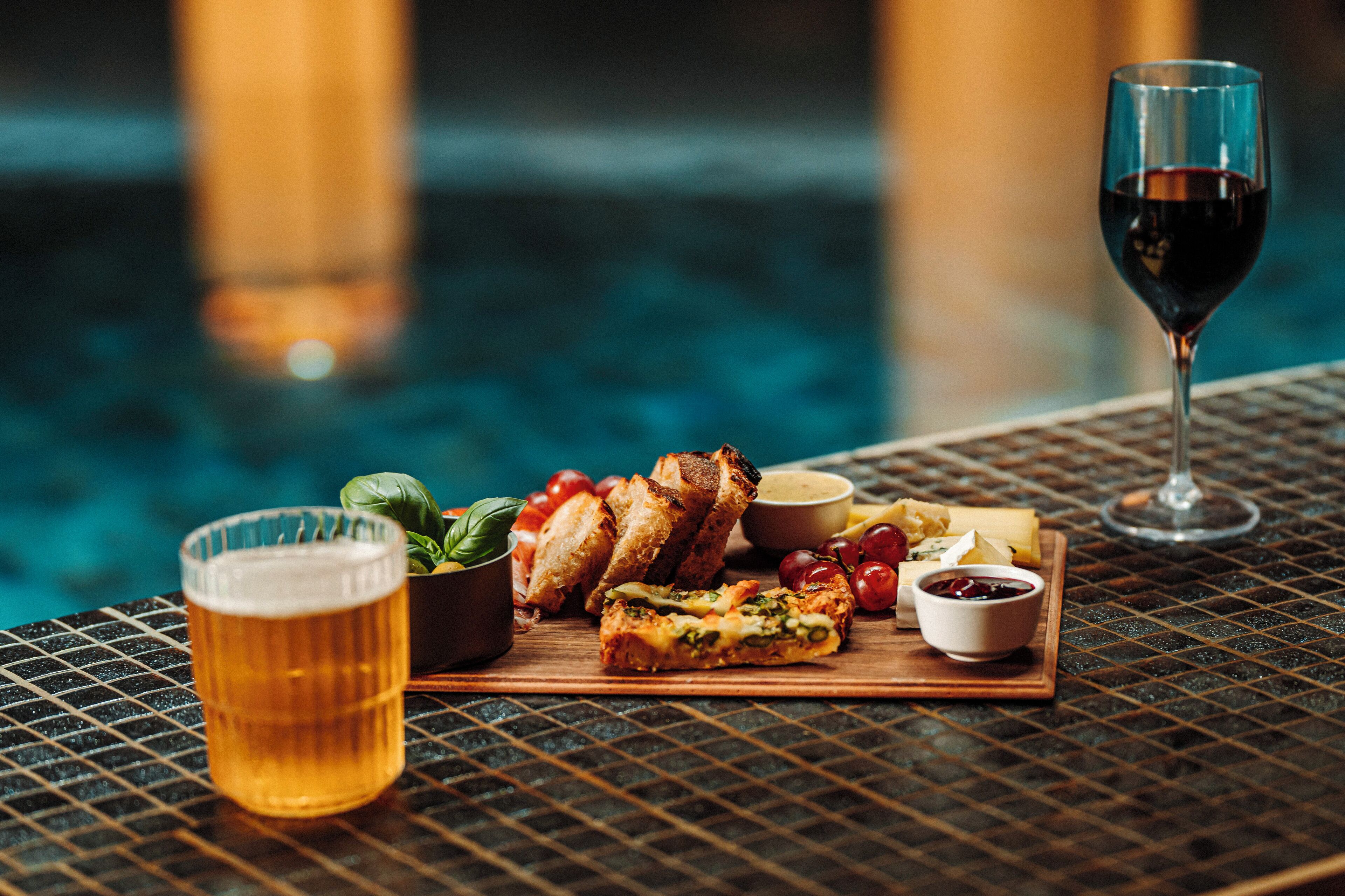 Ein Tisch mit einem Bierglas, einem Weinglas, einem Holzbrett mit gegrilltem Käse, Tomaten und einer Schüssel Soße.