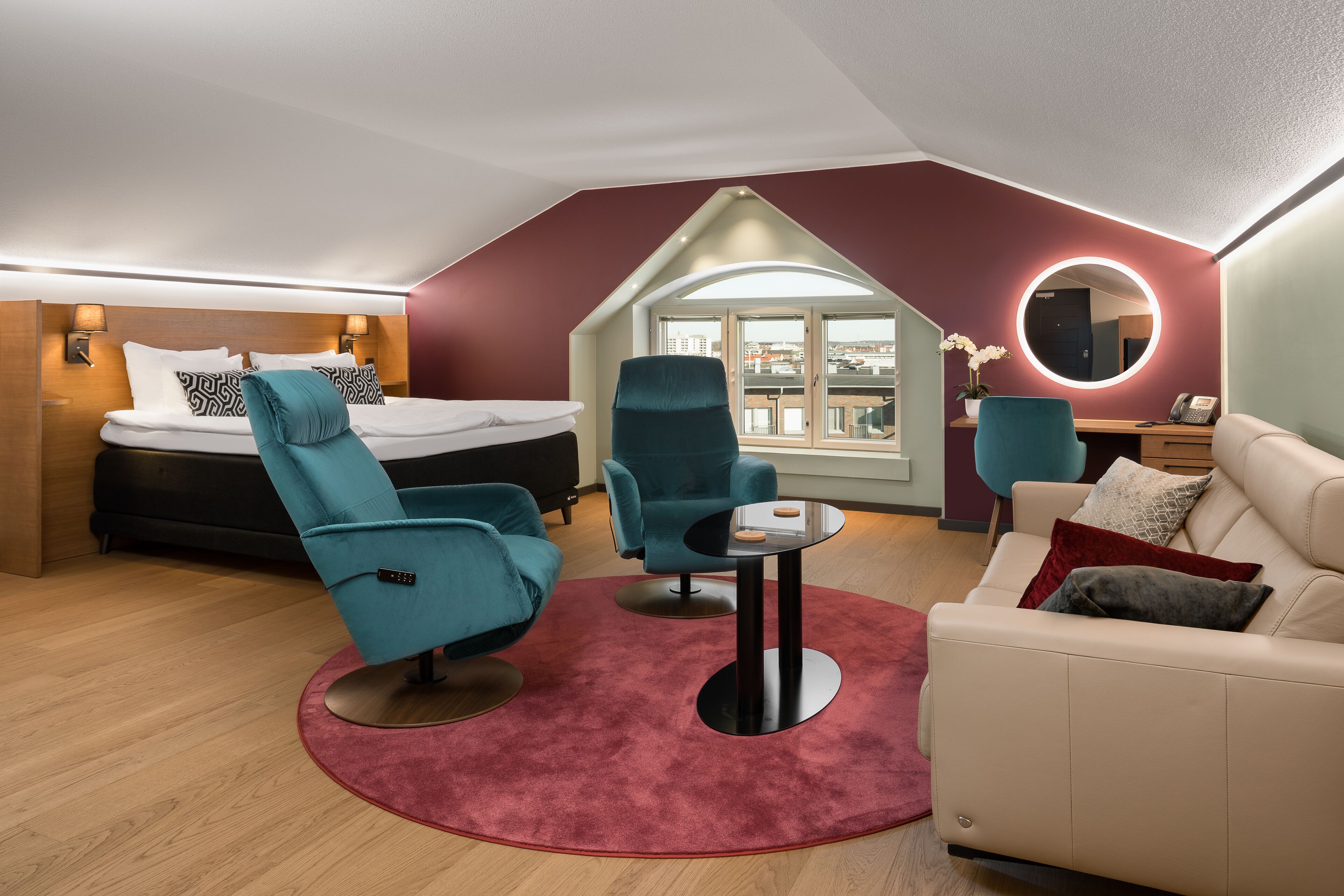 A modern hotel room with a bed, two teal armchairs, a small round table, a sofa, and a large window offering a city view.