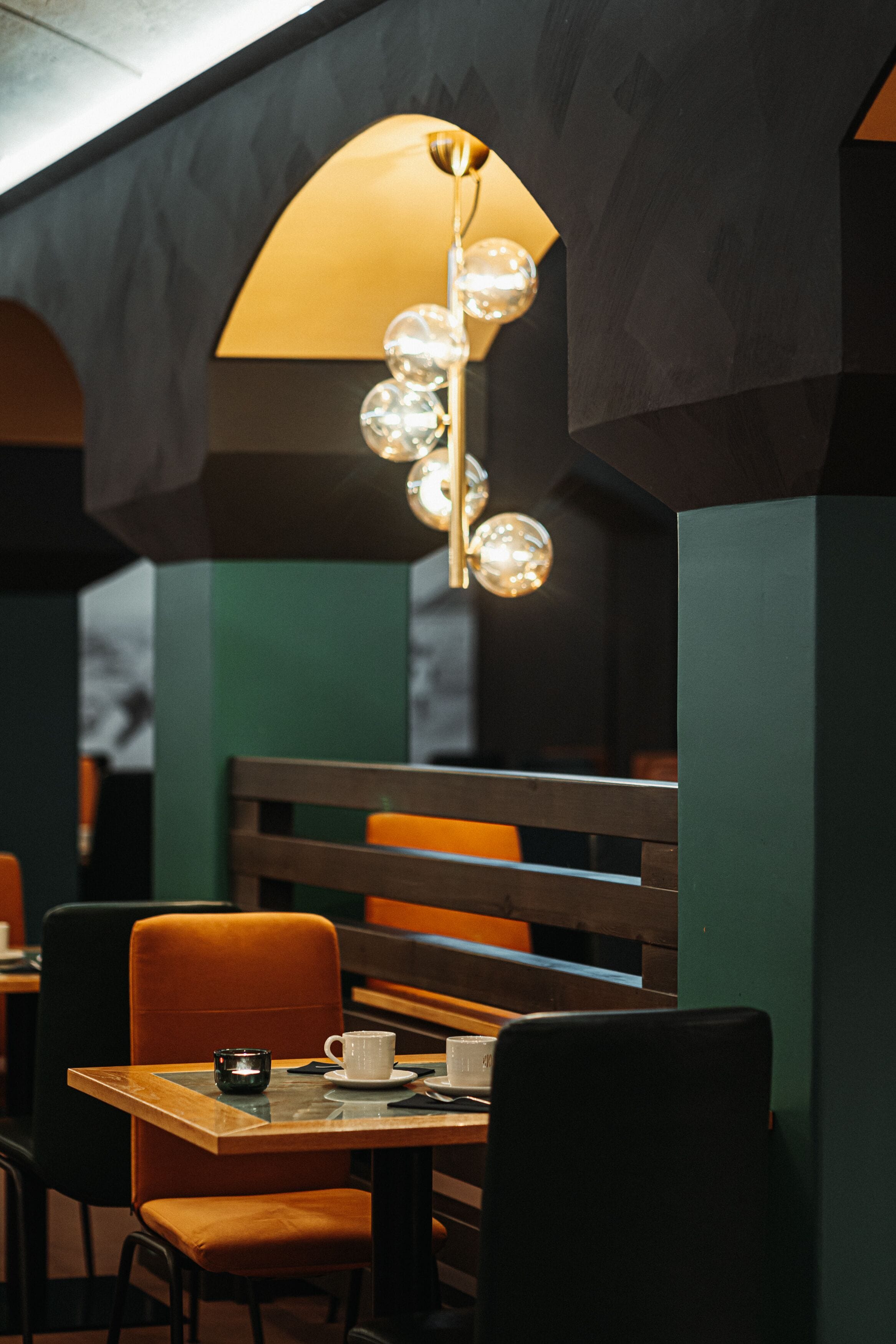 A restaurant interior with a wooden table, orange chairs, and a hanging light fixture with spherical elements.