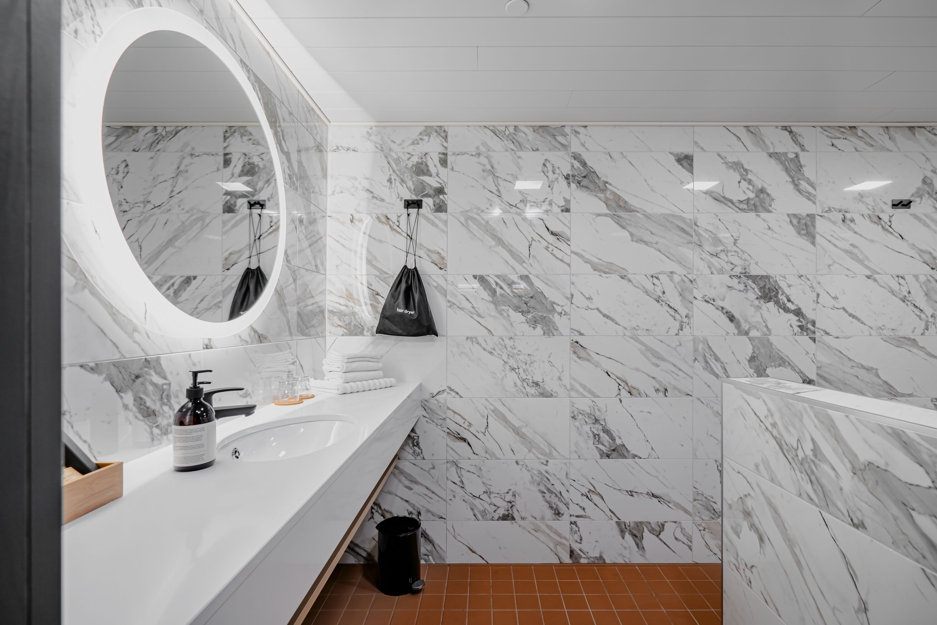 A modern bathroom with a marble backsplash, a large mirror, a white sink, and a black trash bin.