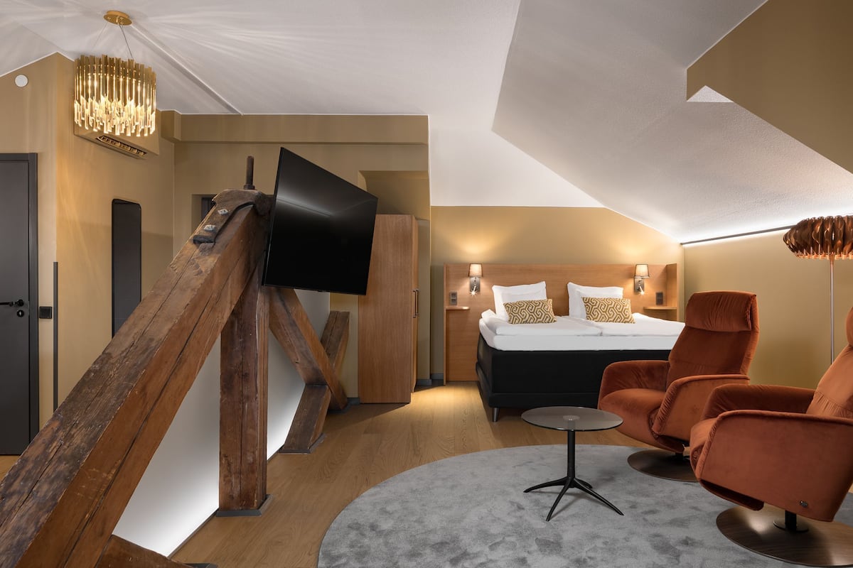 A modern hotel room with a bed, two armchairs, a small table, a TV mounted on a wooden structure, and a chandelier.