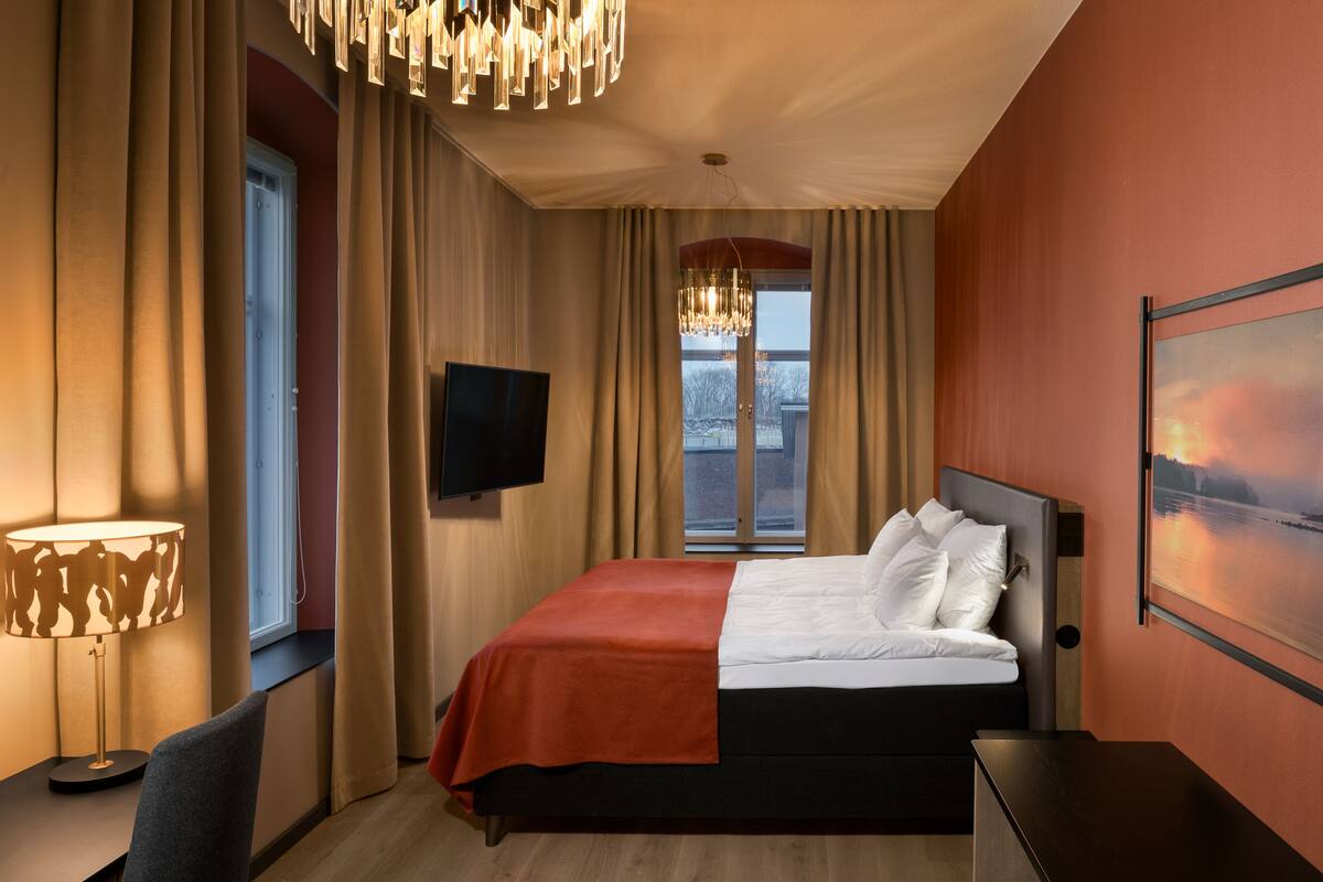 A hotel room with a bed, a TV, a window with curtains, a lamp, and a painting on the wall.