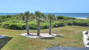 A landscaped area with palm trees and a view of the ocean. - Oceanfront 2BR condo in Caswell Beach, Oak Island—perfect for family getaways! (Oak Island)