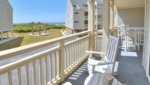 A balcony with white chairs and a table, overlooking a grassy area and buildings. - Oceanfront 2BR condo in Caswell Beach, Oak Island—perfect for family getaways! (Oak Island)