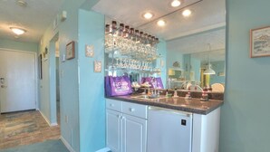 A kitchen with a countertop, a refrigerator, and a shelf with glassware. - Oceanfront 2BR condo in Caswell Beach, Oak Island—perfect for family getaways! (Oak Island)