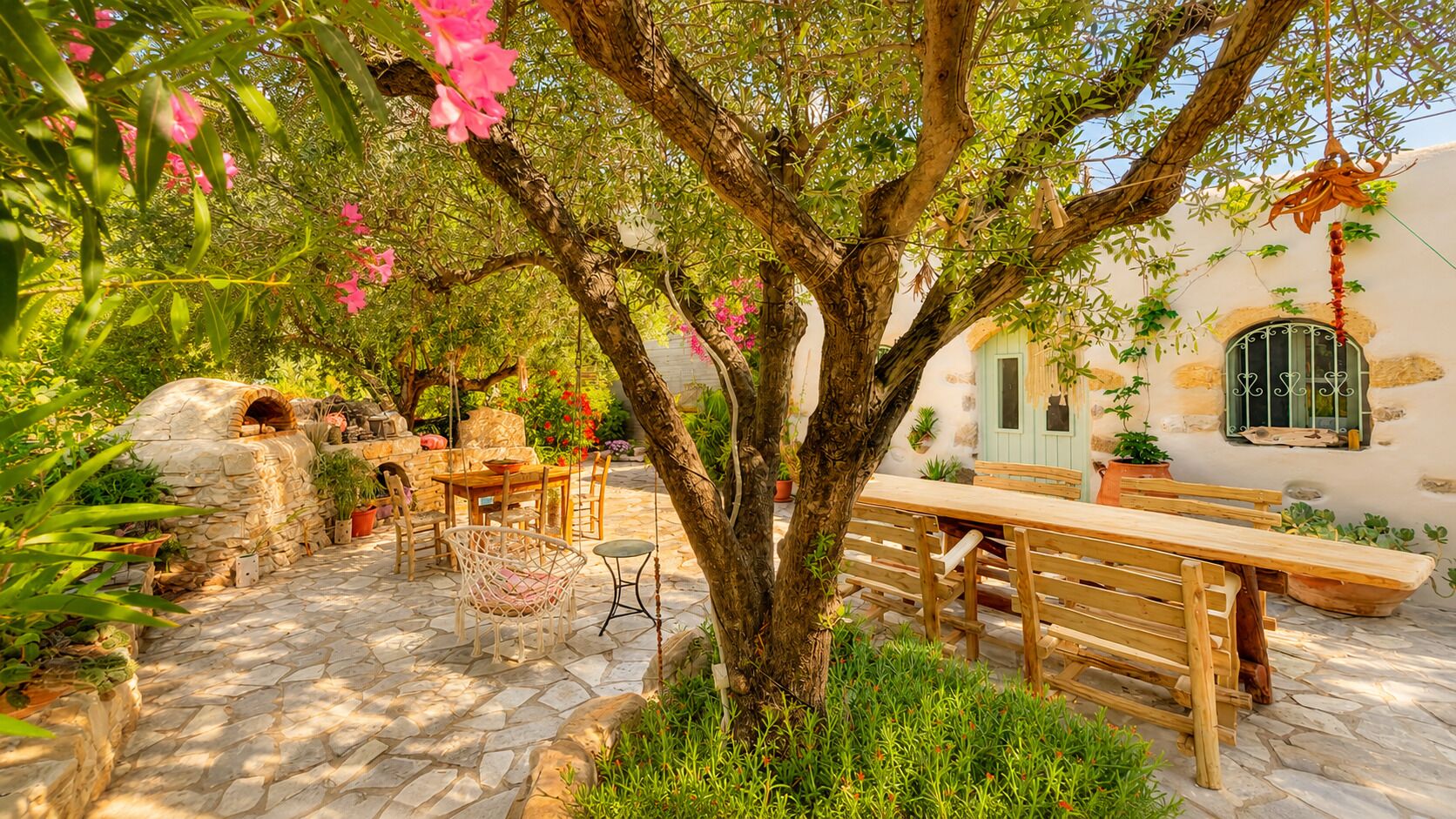 Un patio empedrado con una mesa y sillas de madera, un horno de piedra y un árbol con flores rosadas.