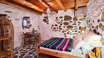 Une pièce rustique avec un mur en pierre, un plafond en bois, un lit recouvert d’une couverture rayée aux couleurs vives, une chaise en osier et une petite table avec une lampe.