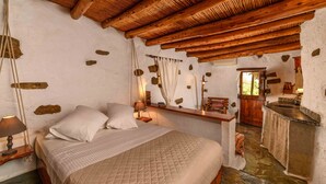 A rustic room with a wooden ceiling, a bed with white bedding, a small desk with a lamp, and a stone wall. - Natura cottages (Sitia)