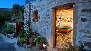 A rustic stone building with a wooden bed and a stone floor. There are potted plants outside and a small window with a plant inside. - Natura cottages (Sitia)