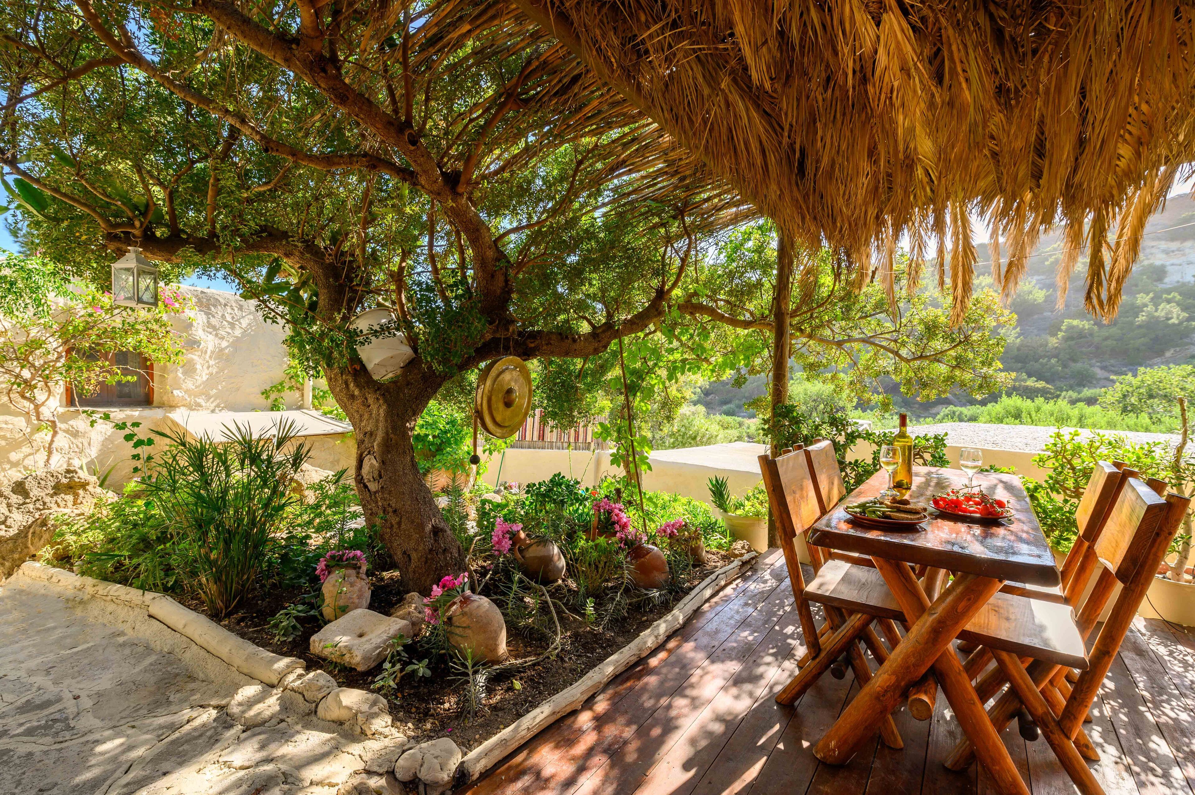 An outdoor dining area with a wooden table and chairs, surrounded by lush greenery and a tree.