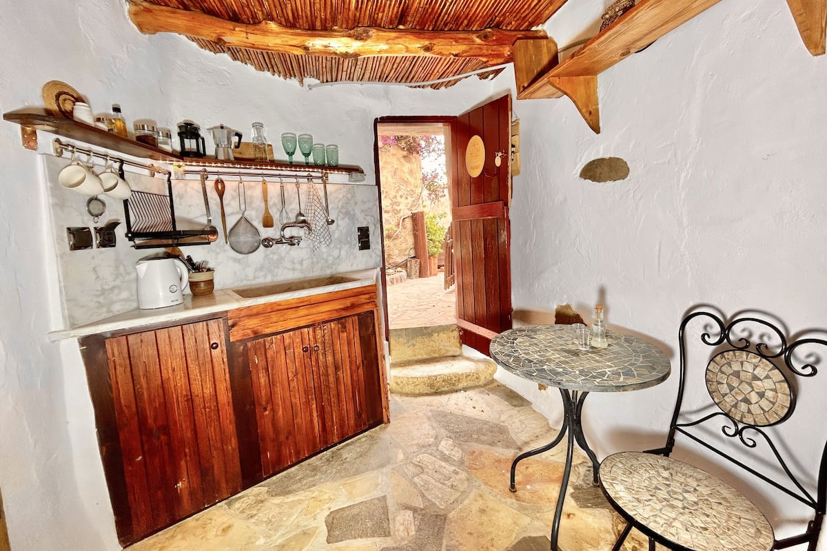 Una cocina rústica con armarios de madera, suelo de piedra y un pequeño comedor con una mesa y sillas de metal.