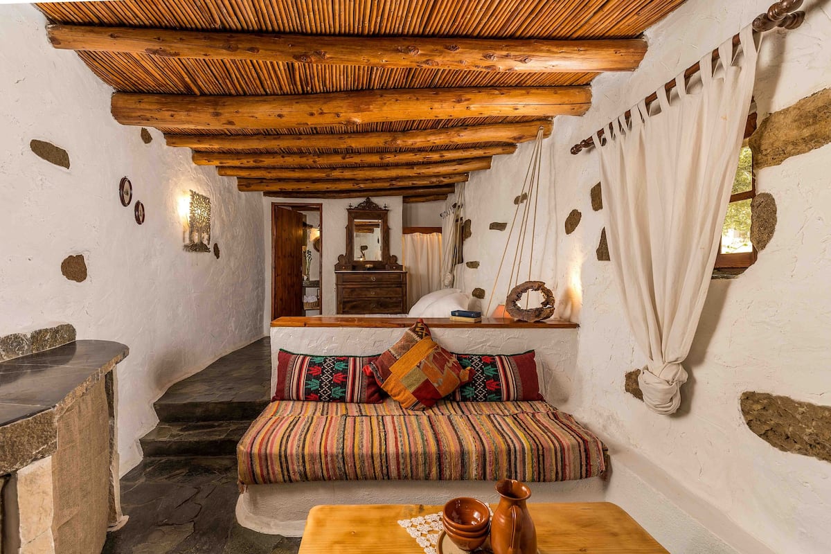 Un interior rústico con techo de madera, paredes de piedra, un banco con cojines de colores y una mesa de piedra con cerámica.