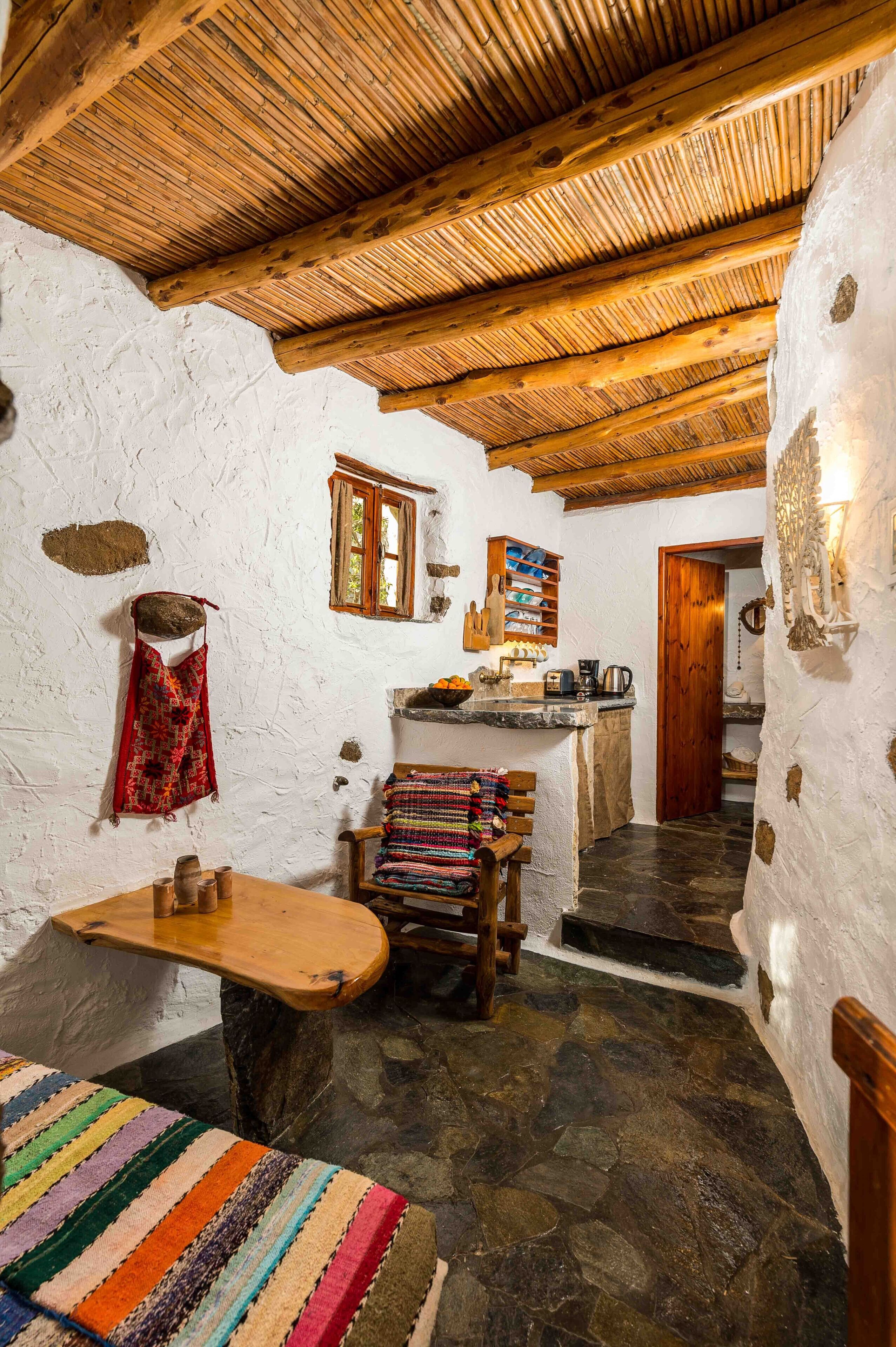 Un interior rústico con suelo de piedra, vigas de madera, un banco, una mesa y una alfombra colorida.