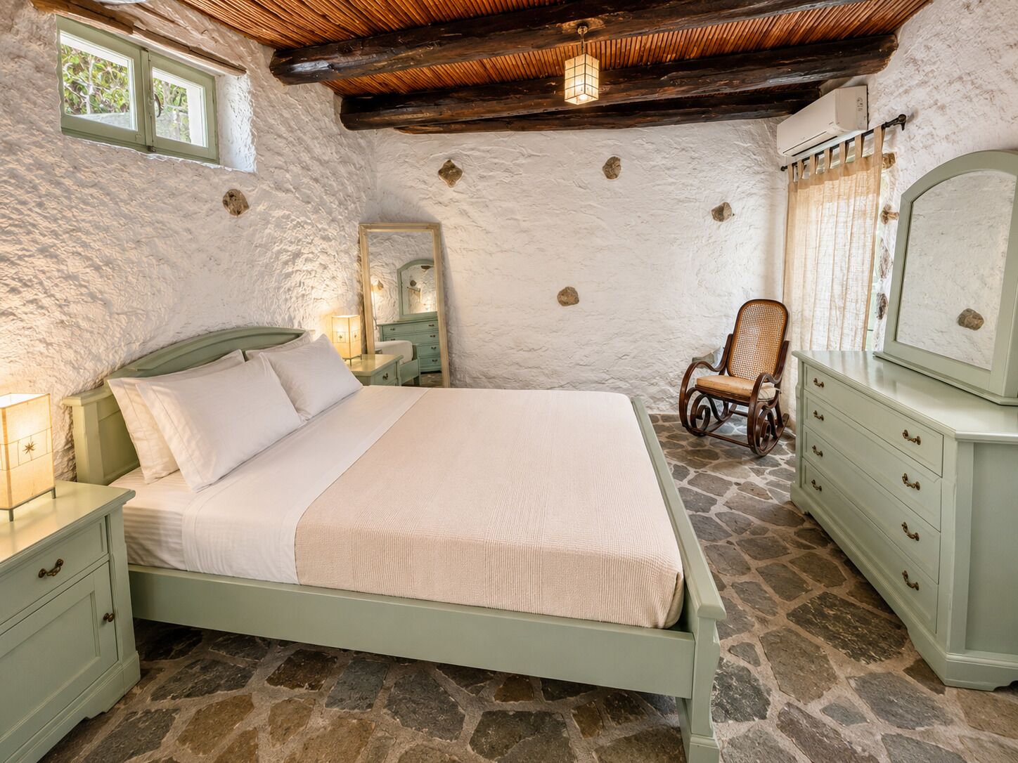 A bedroom with a stone floor, a bed with white linens, a dresser, a chair, and a mirror.