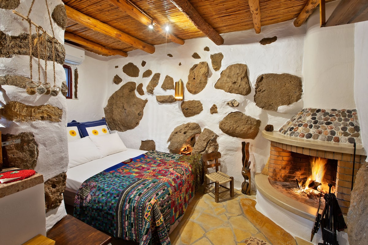 Una habitación rústica con pared de piedra, una cama con colcha de colores, chimenea y techo de madera.