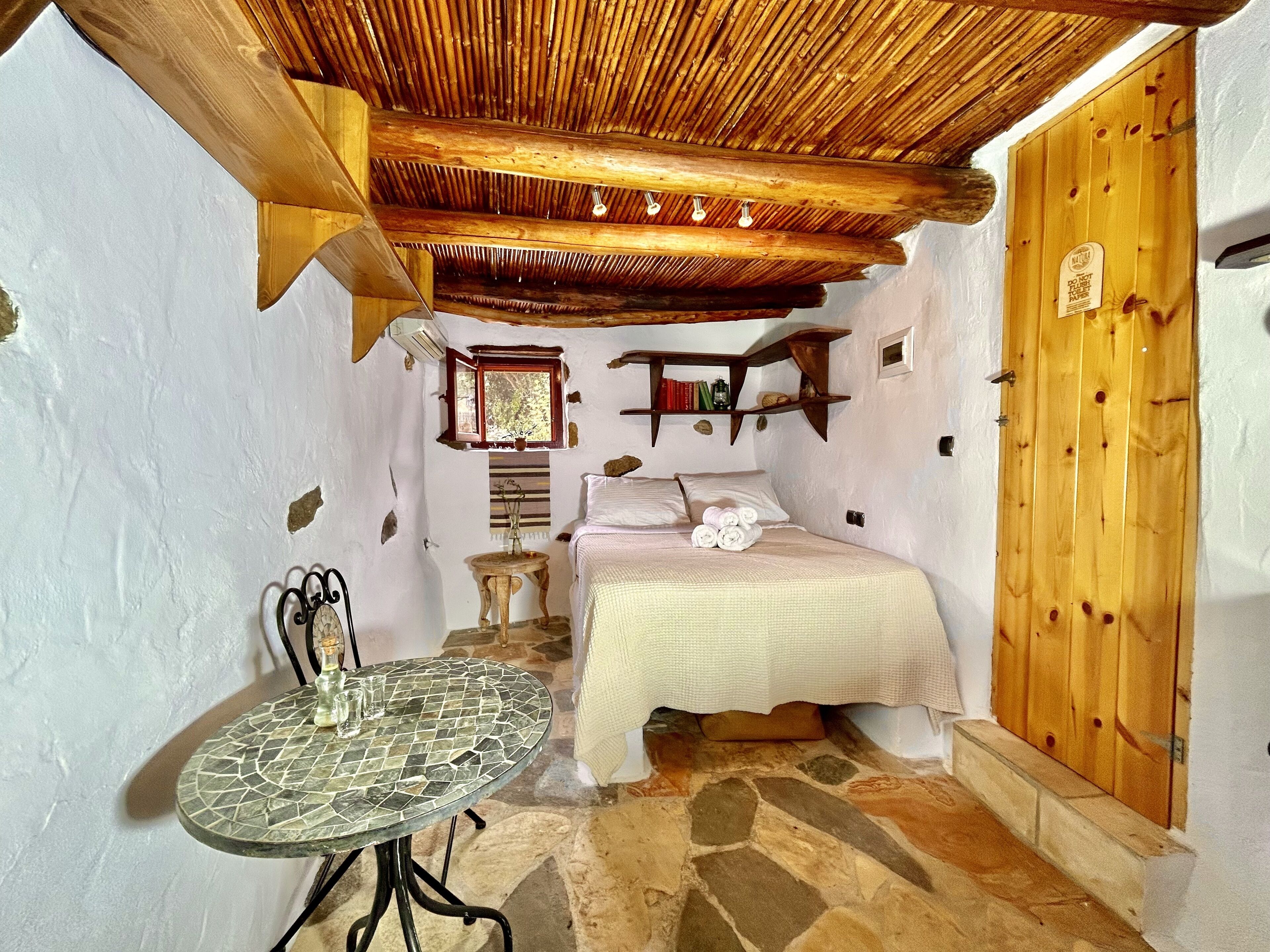 Quarto rústico com cama, uma mesinha, uma estante com livros e uma porta de madeira.