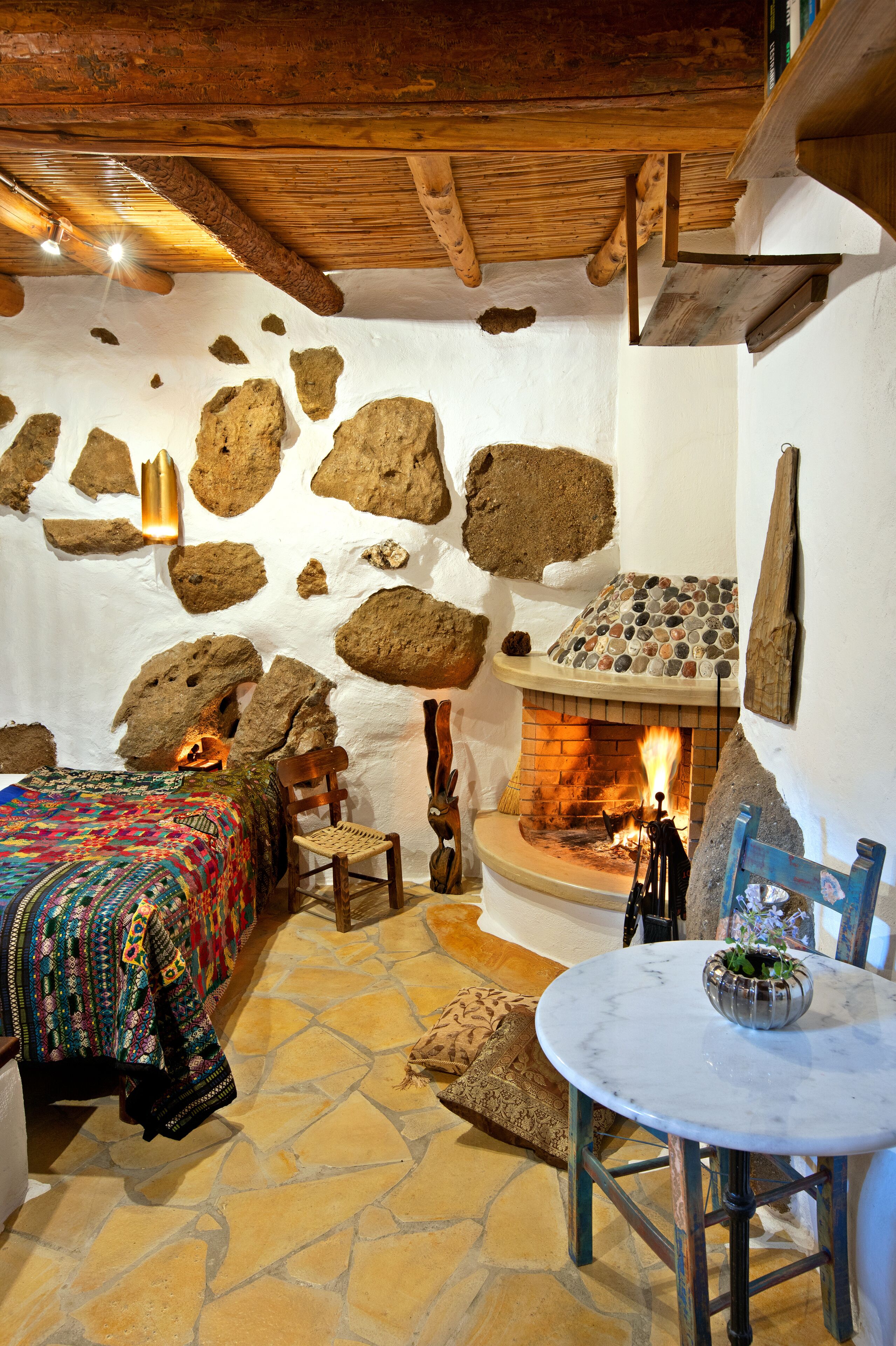 Una habitación rústica con pared de piedra, chimenea, cama con manta de colores y una mesa redonda con un cuenco.