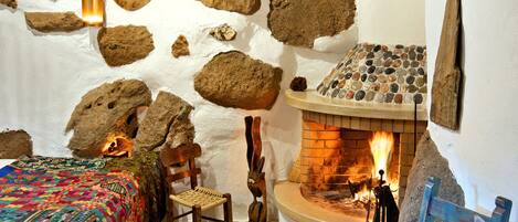 Una habitación rústica con pared de piedra, chimenea, cama con manta de colores y una mesa redonda con un cuenco.