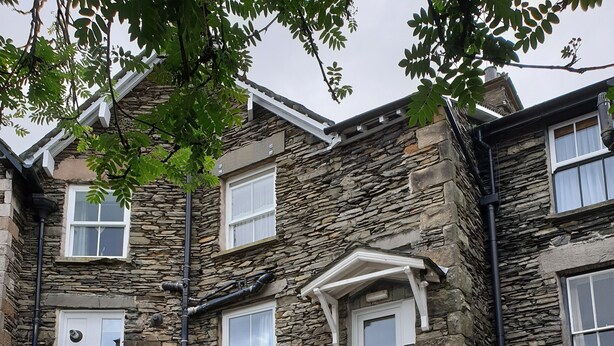 A stone building with multiple windows, a front entrance with a door, a small porch with a bench, and a staircase leading to an upper floor.