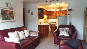 A living room with a red leather sofa and armchair, a framed picture on the wall, and a kitchen in the background.