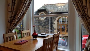 A dining area with a wooden table, chairs, a vase with a flower, and a view of a stone building through a large window.