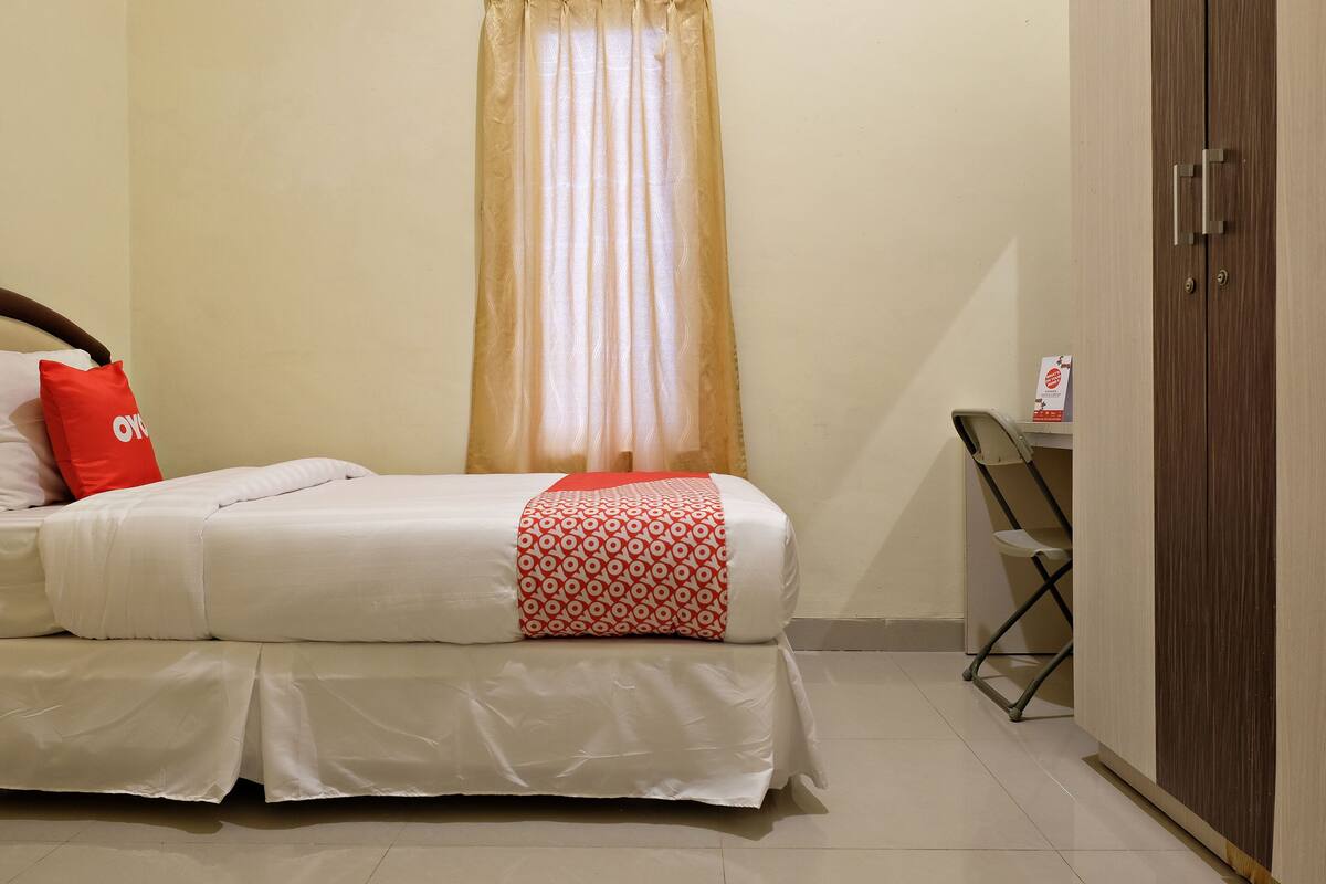 Un lit bien fait, recouvert d’une couverture à motifs blancs et rouges, avec un oreiller rouge et un rideau beige.