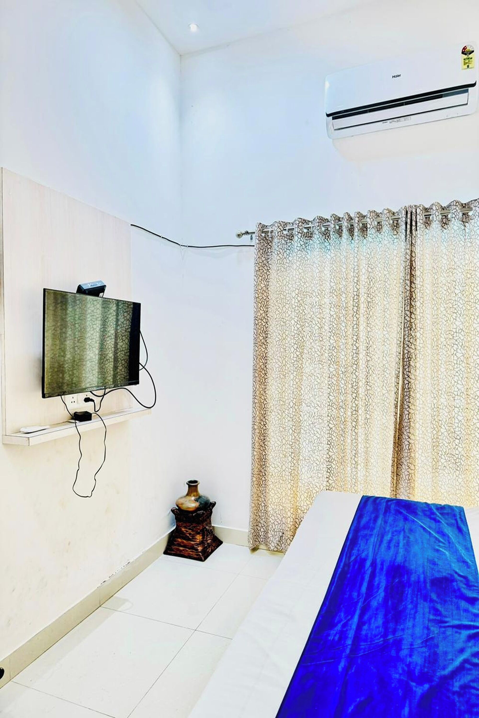 A bedroom with a bed, a television mounted on the wall, a small table with a vase, and a curtain.