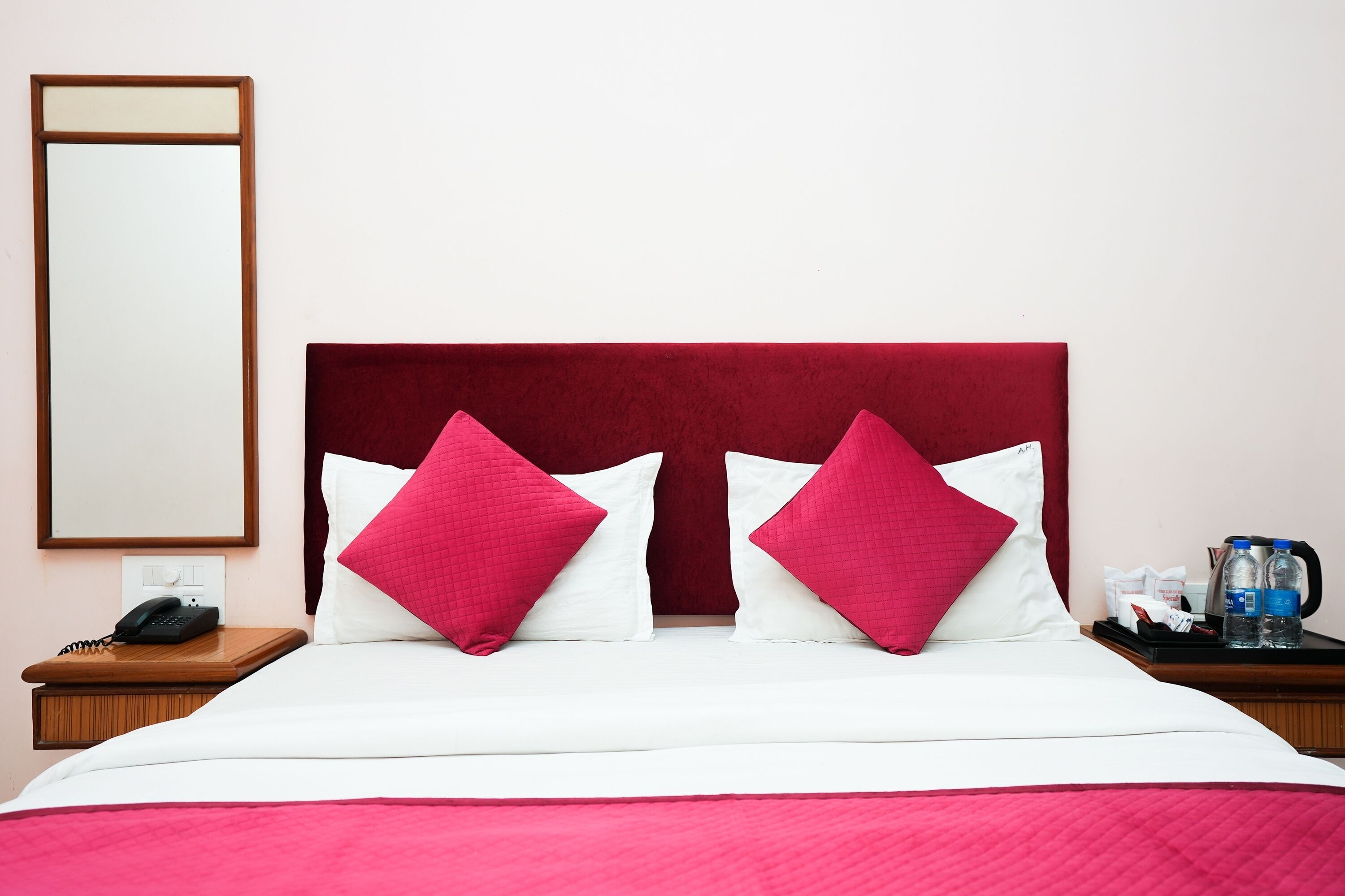 A hotel room with a bed, two red pillows, a white bedspread, a wooden nightstand with a telephone, and a small table with water bottles and a coffee pot.