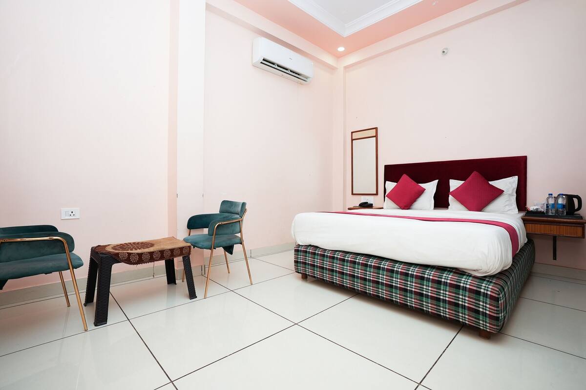 A hotel room with a bed, a small table, two chairs, a nightstand, and an air conditioning unit.