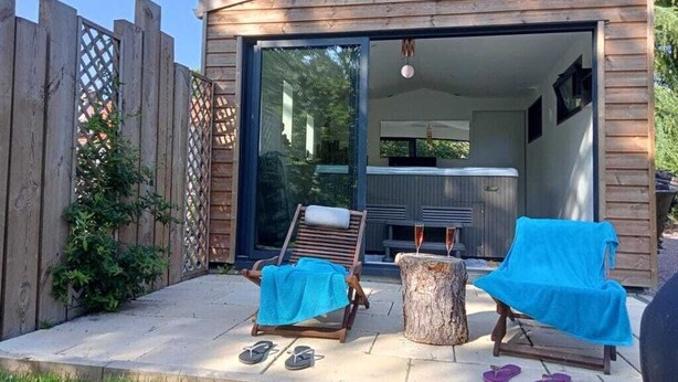 A wooden cabin with a sliding glass door leading to a room with a bed and a heater. There are two lounge chairs with towels, a tree stump, and a pair of flip-flops on the patio. - Logis du Parc (Saint-Fulgent)