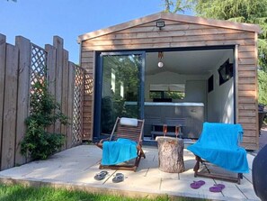 A wooden cabin with a sliding glass door leading to a room with a bed and a heater. There are two lounge chairs with towels, a tree stump, and a pair of flip-flops on the patio.