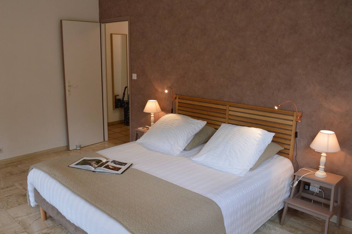 Un lit bien fait, avec une couverture beige et des oreillers, deux lampes de chevet et un magazine posé sur le lit.