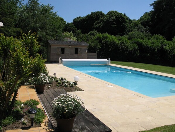 A swimming pool with a wooden deck, surrounded by lush greenery and a small building in the background.