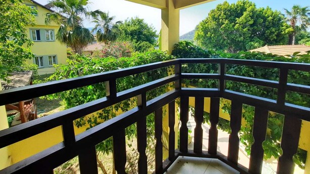 A balcony with a wooden railing overlooking a garden with various plants and a yellow building in the background. - Dalyan Summer Life Apart Hotel (Ortaca)