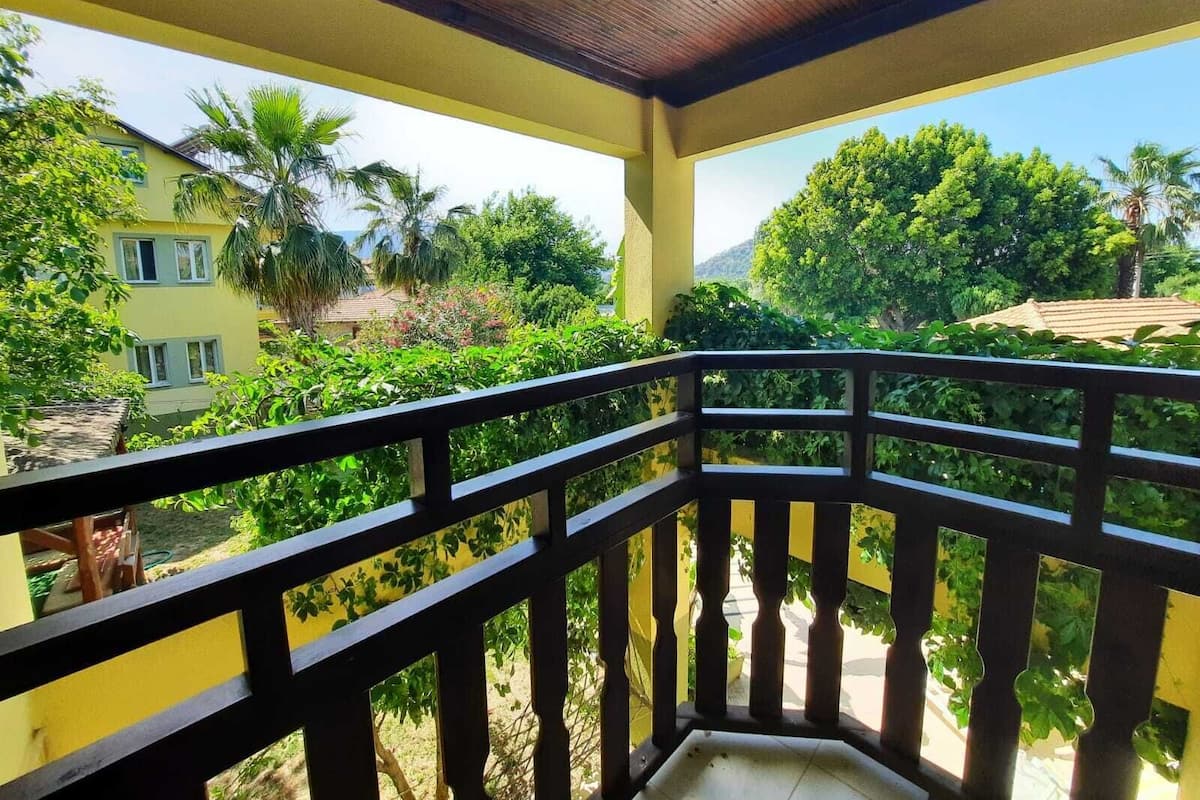 A balcony with a wooden railing overlooking a garden with various plants and a yellow building in the background.