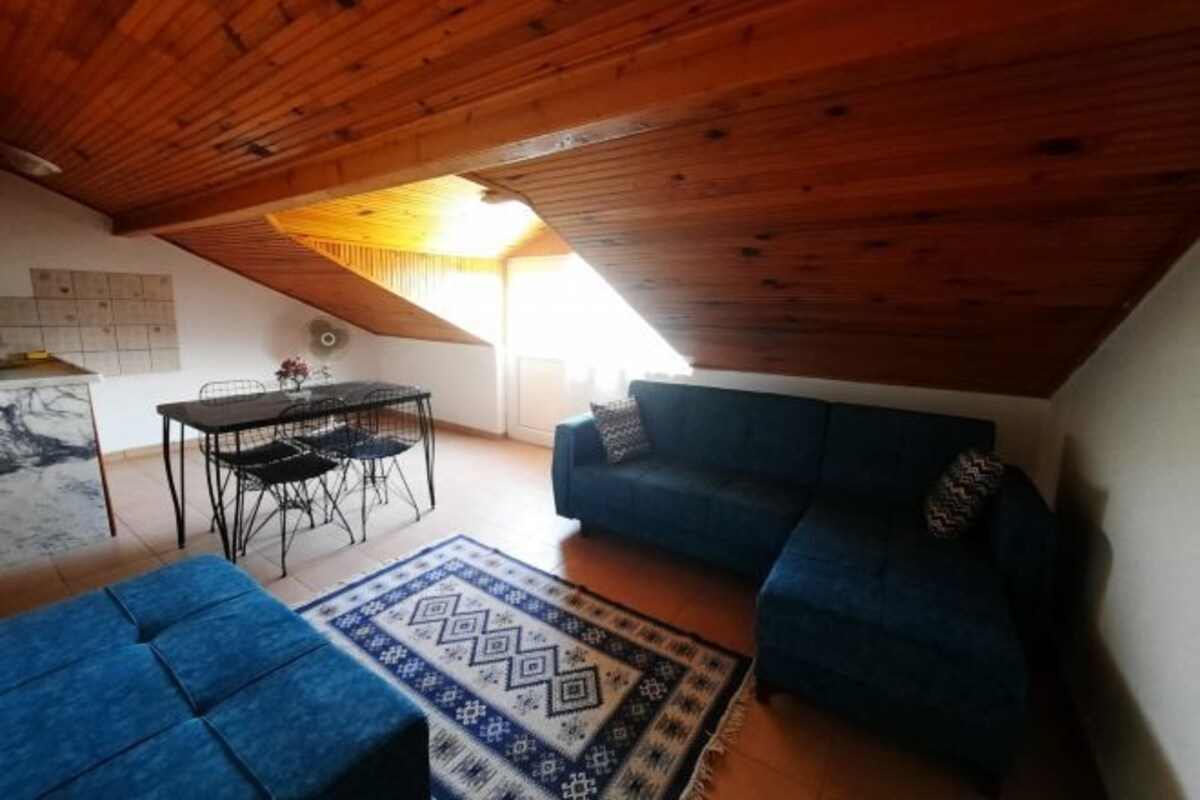 A cozy loft-style living space with a wooden ceiling, a blue sofa, a dining table with chairs, and a patterned rug.