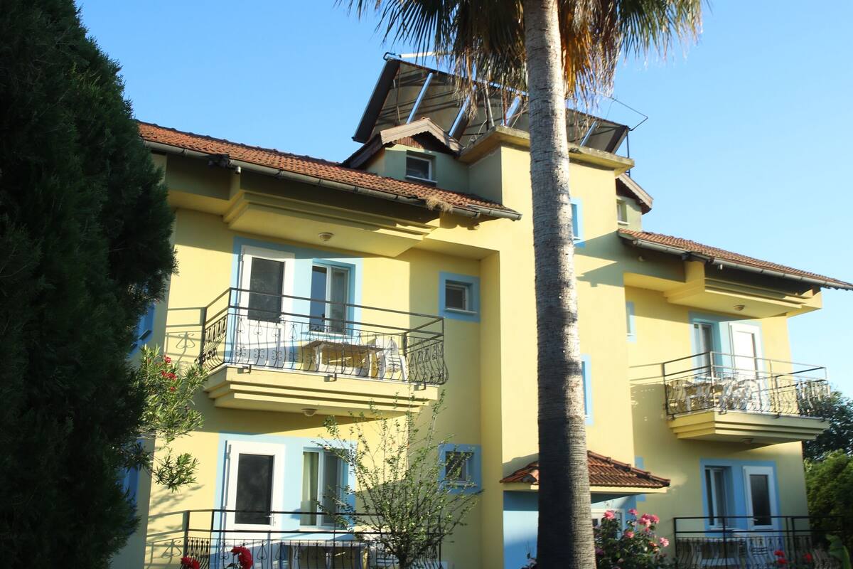 A two-story building with balconies, a palm tree, and red flowers in the garden.
