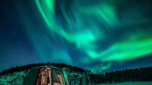 Une structure transparente en forme d’igloo, dont l’intérieur est visible, installée sur un paysage enneigé sous un ciel nocturne illuminé par les aurores boréales.